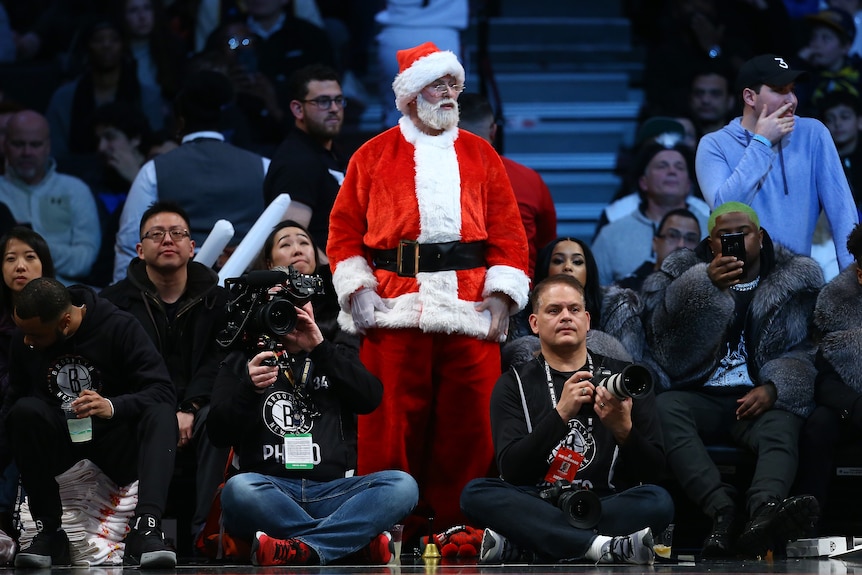 A man dressed as Santa Claus stands with his arms by his side next to other people who are sitting