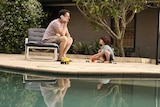 Man and boy sit inside backyard pool fence. Boy plays with trucks while dad is distracted on the phone