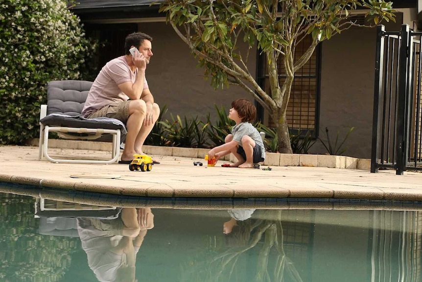 Man and boy sit inside backyard pool fence. Boy plays with trucks while dad is distracted on the phone