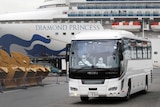 Drivers with protective suits drive a bus next to earth movers with a cruise ship in the background
