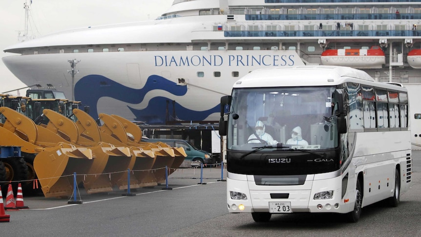 Drivers with protective suits drive a bus next to earth movers with a cruise ship in the background