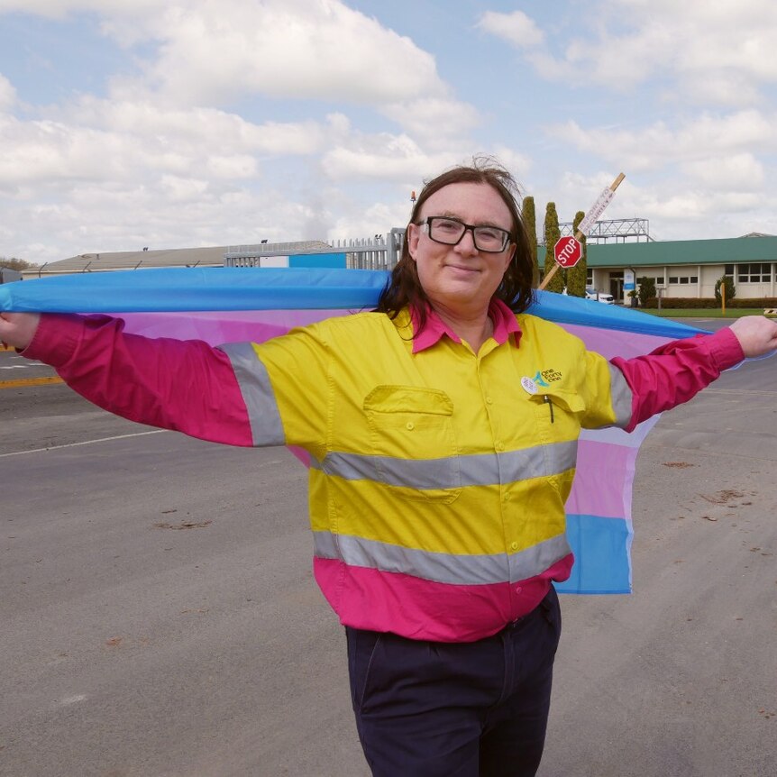 A woman smiles at the camera, holding a blue, pink, and white striped flag.