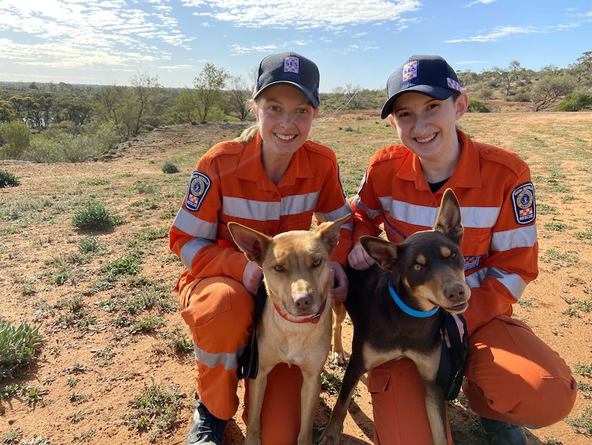 Beatrice Walladge and Sophie Western in orange SES overalls with two kelpies and scrub and river in background.