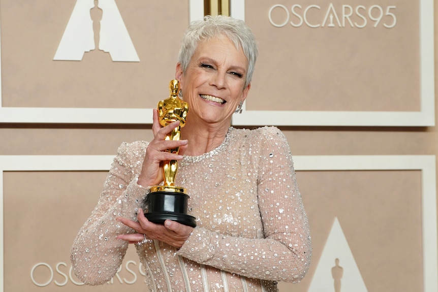 A slim white woman in her 60s with short white hair beams as she holds a golden statuette up in front of an Oscars backdrop.