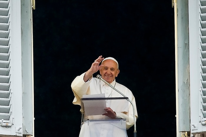 A man in a white outfit waves with his right hand from a large open window while smiling