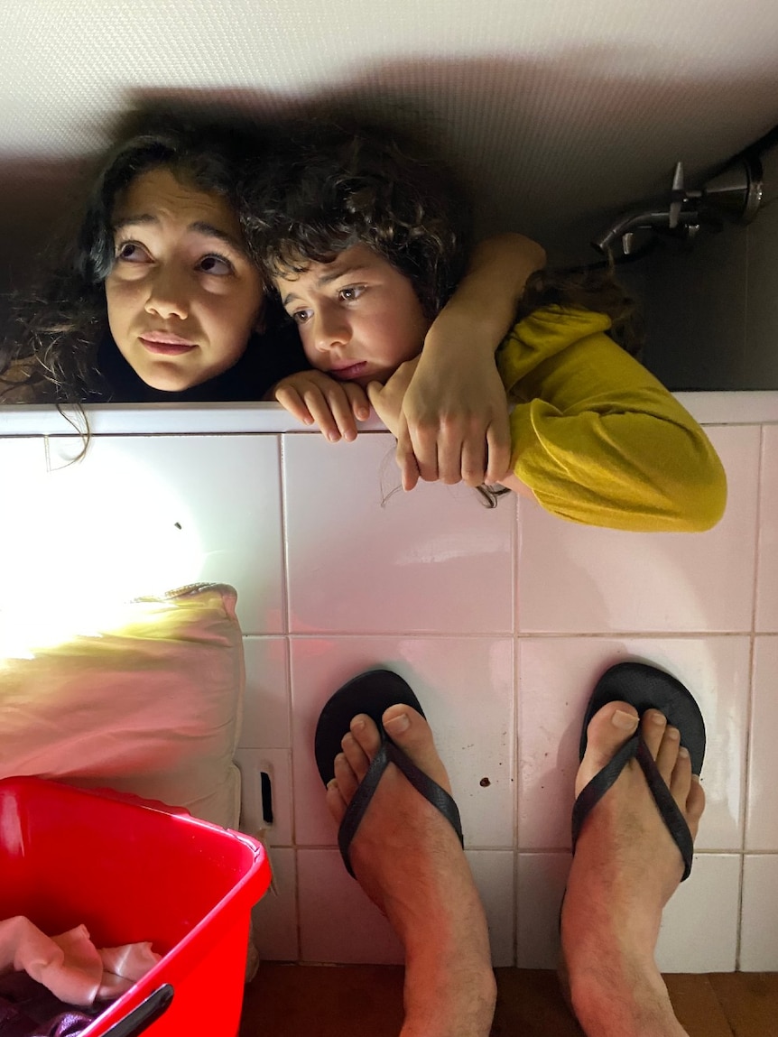 Two girls looking frightened sheltering under mattress in a bath.