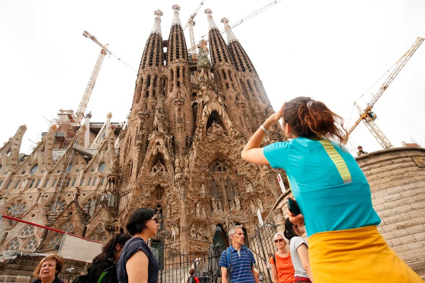 A person shields their face from the sun as they look up into the ornate spires of La Sagrada Familia church.