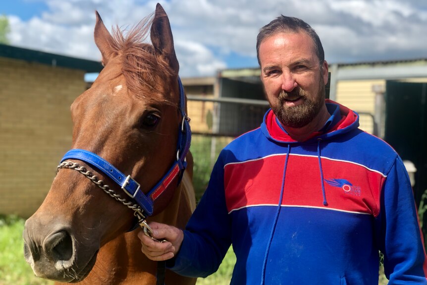 A bearded man looks at the camera as he holds a brown horse by its bridle.