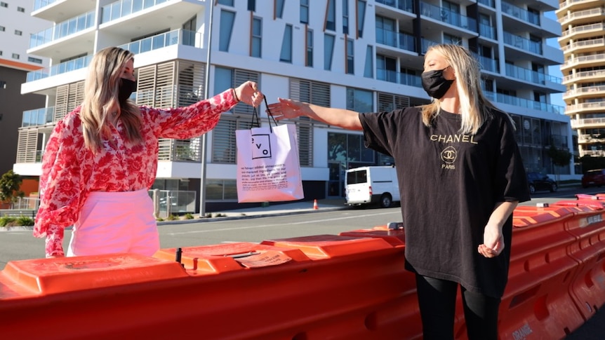 Lady in black stands on one side of barricade and hands shopping bag to lady in pink