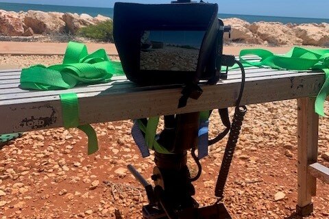 Camera upside down, attached to a bench, with red dirt and beach in background.
