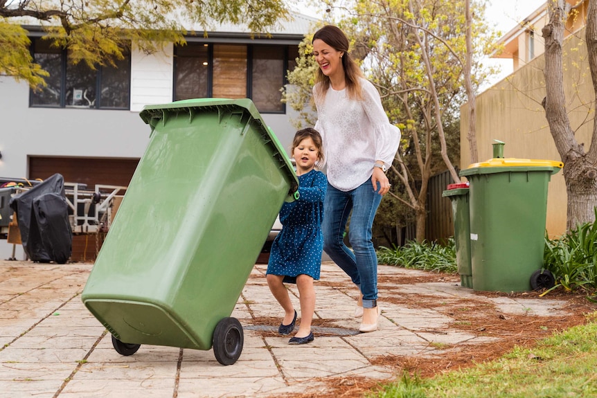 A small girl in a blue dress and a woman wearing jeans push a green bin on a driveway.