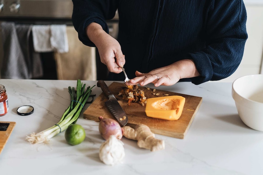 A chopping board with butternut pumpkin and a knife, surrounded by spring onions, a shallot, ginger, garlic, for a soup.