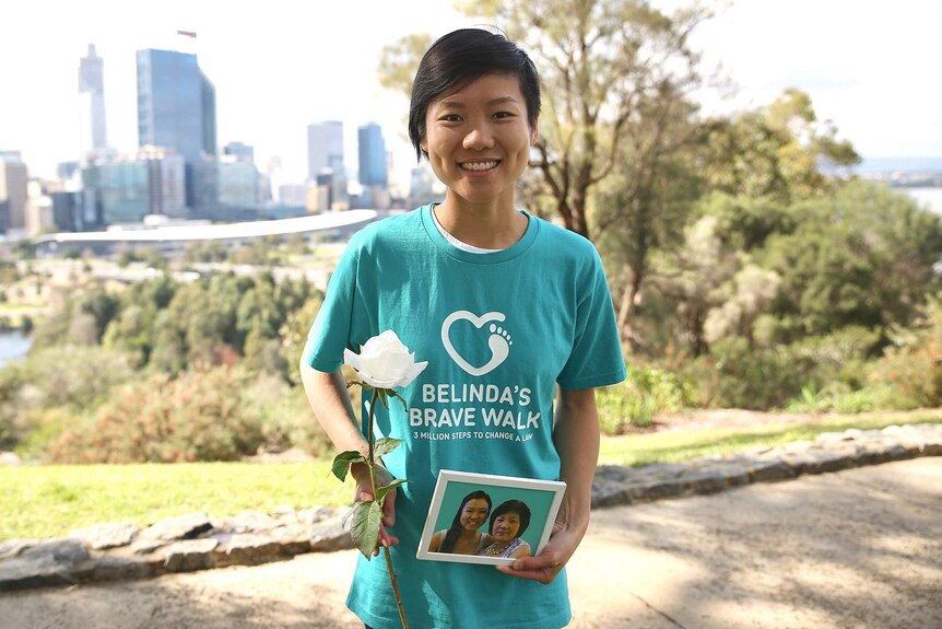 Belinda Teh poses for a photo at Kings Park in Perth holding a white rose and a photo while smiling.