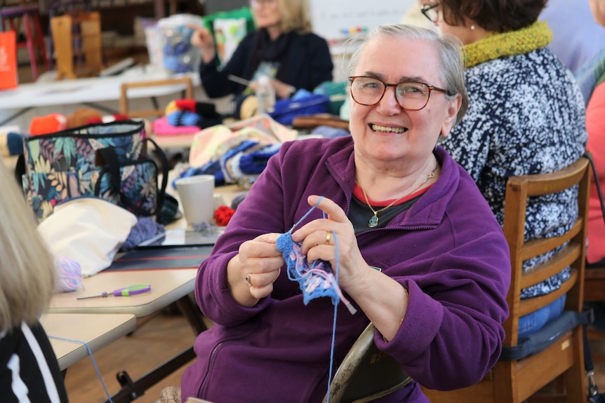 A woman sitting down looks at the camera smiling while knitting