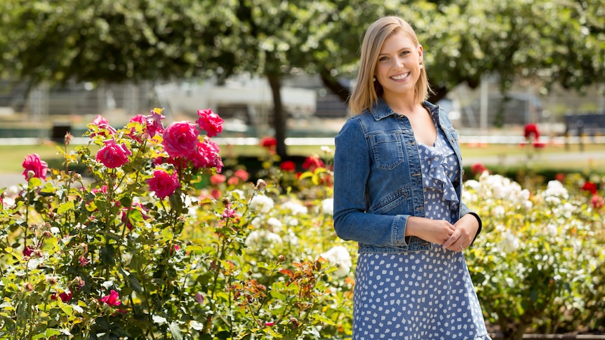 A woman with shoulder-length blonde hair, wearing a blue dress and denim jacket, standing by a rose garden.
