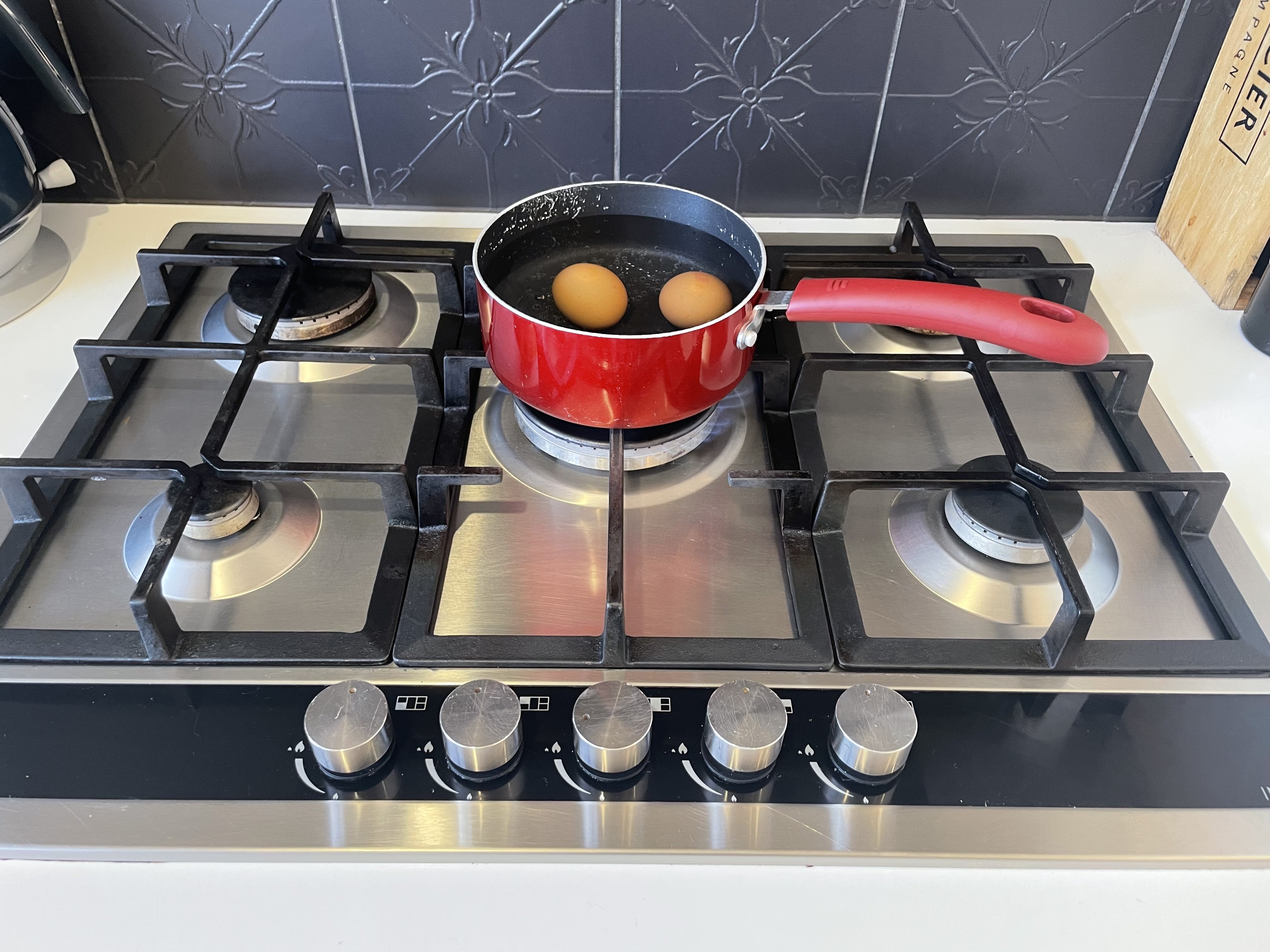 Two eggs boil in water in a red saucepan on a stainless steel gas cooktop with a black patterned tile splashback