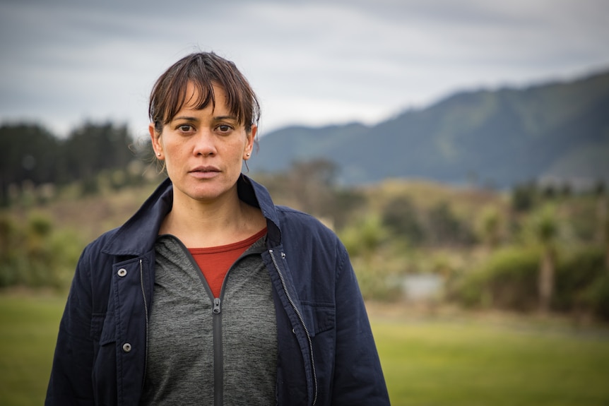 A scared-looking Maori woman in her 40s stands in the New Zealand countryside, dressed in activewear
