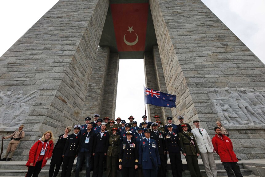 A group of New Zealand officials in uniforms wearing medals hold a New Zealand flag with a Turkish flag hovering above them.
