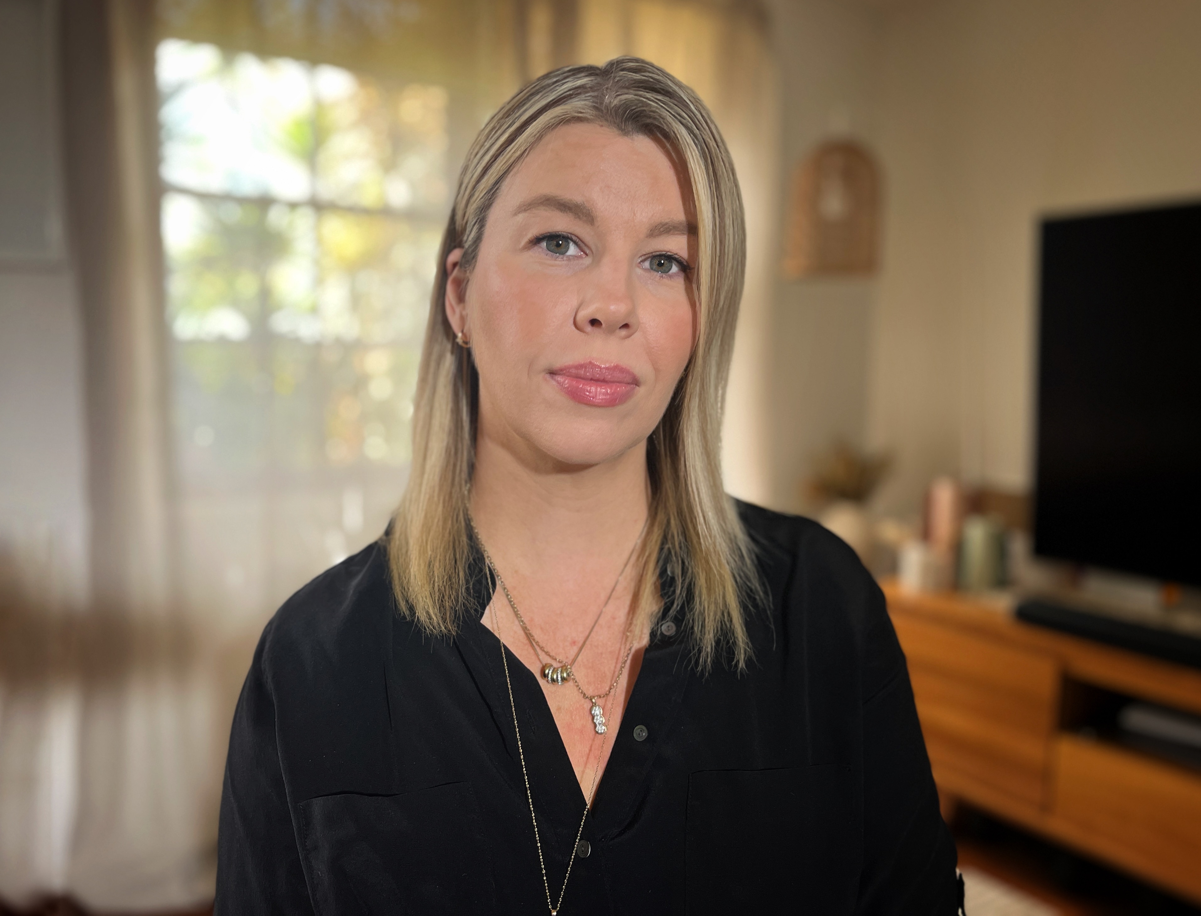 A blonde, long-haired woman (Gemma Wilson) wearing a black blouse sits in a lounge room setting.