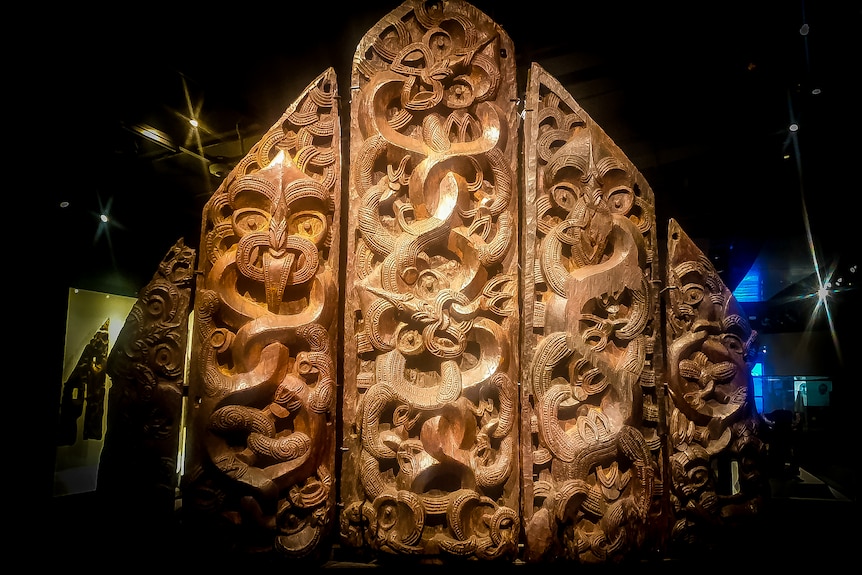 Five intricately carved wooden panels spotlit in a dark room. 