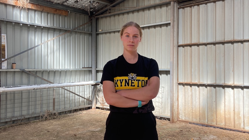A woman, arms crossed and with a serious expression, stands in a large empty shed with a dirt floor. 