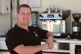 A short haired man wearing a black polo stands in front of a coffee machine