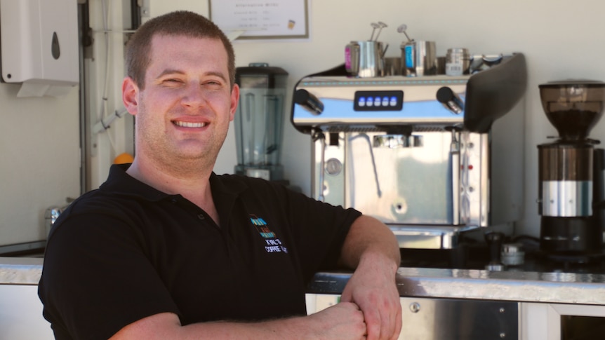 A short haired man wearing a black polo stands in front of a coffee machine