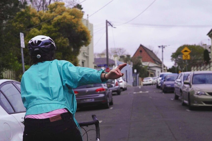 Julia is riding her bike on the left hand side of the road, she is reaching out with her right hand to indicate a right turn.