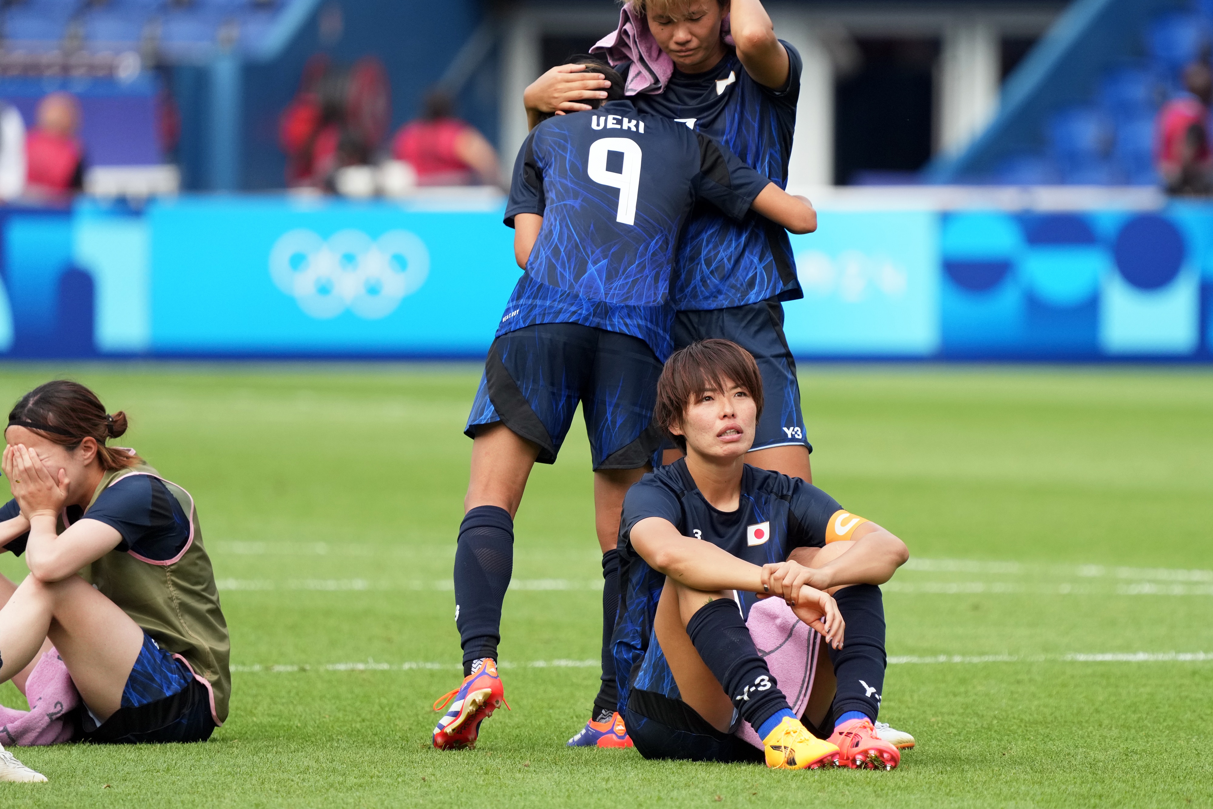 Uma jogadora está sentada em campo e parece desanimada enquanto seus companheiros se abraçam.