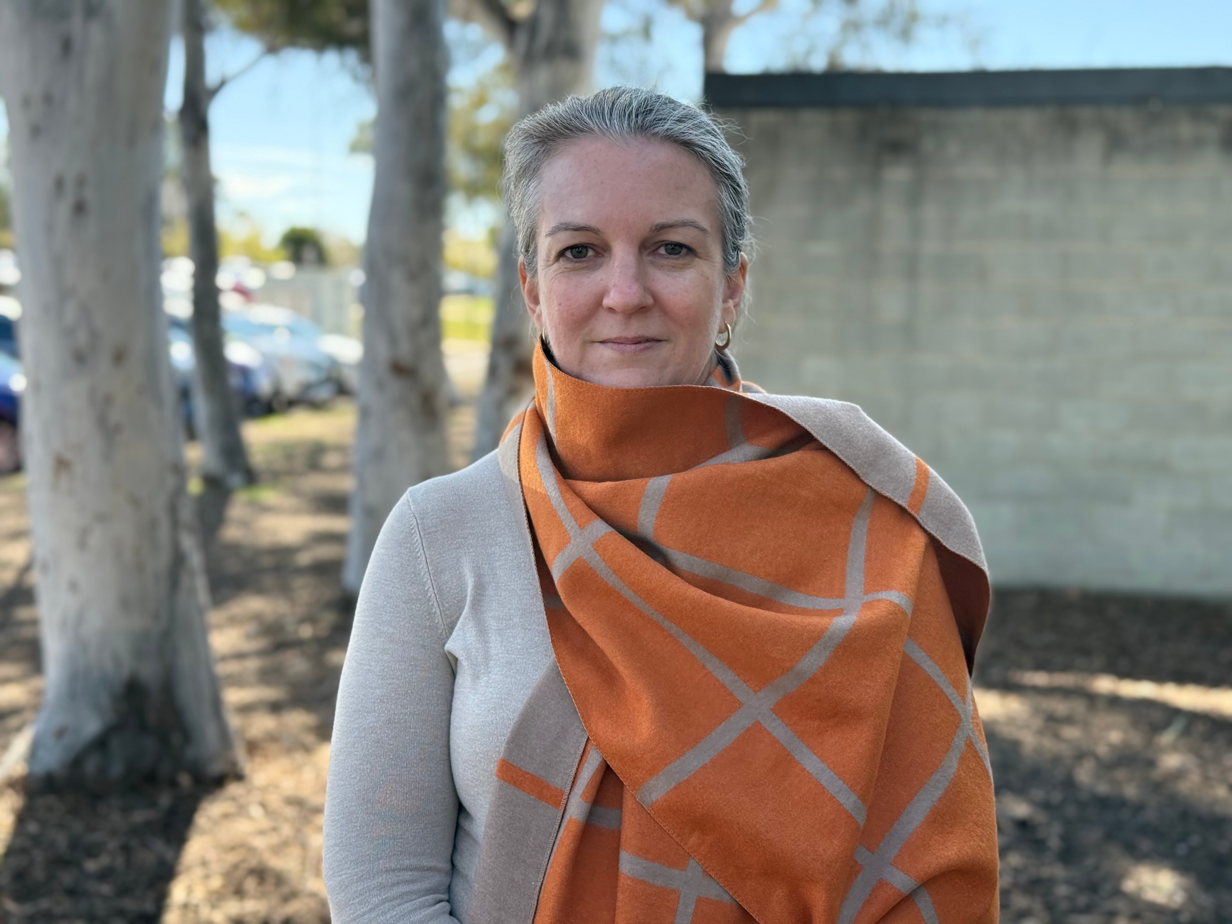 A middle-aged woman with grey hair tied back wearing a large orange scarf stands outside smiling.