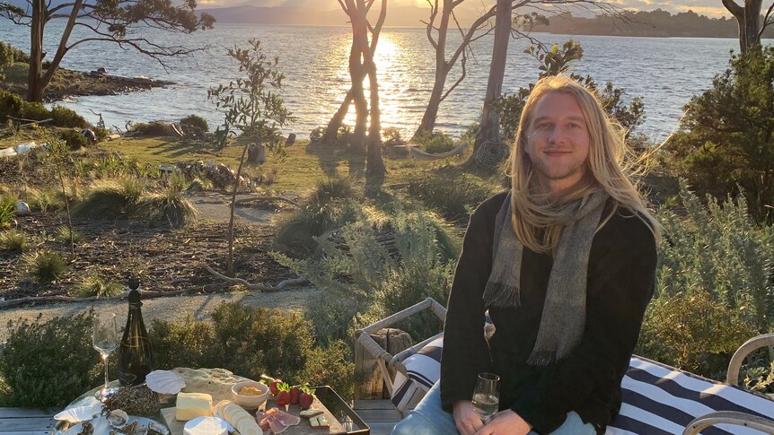 A young man with long blonde hair is setting on a bench with a glass of wine in his hands and a cheeseboard next to him.