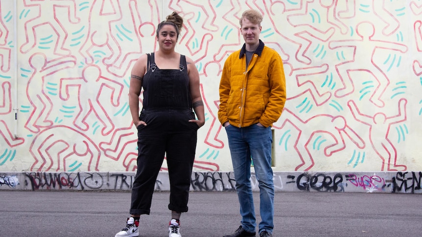 A young woman and a young man looking at the camera and standing in front of a wall daubed with street art.