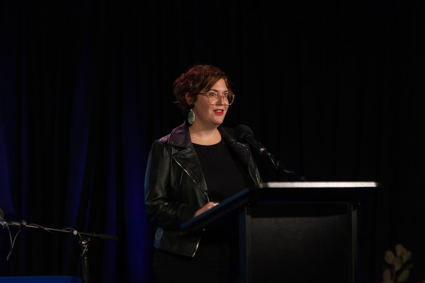 A middle-aged brunette woman with short hair and glasses, wearing a leather jacket, stands at a podium on a stage 