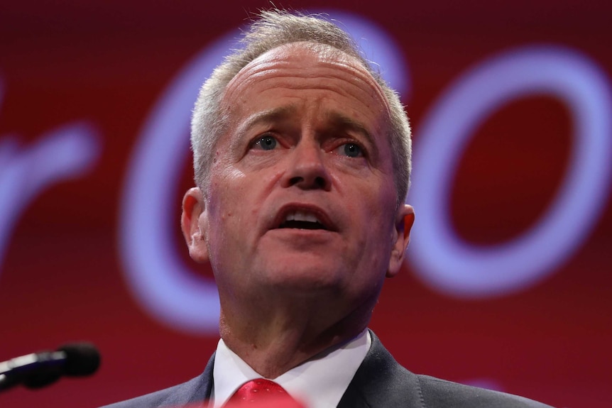Bill Shorten speaks behind a microphone with a red background