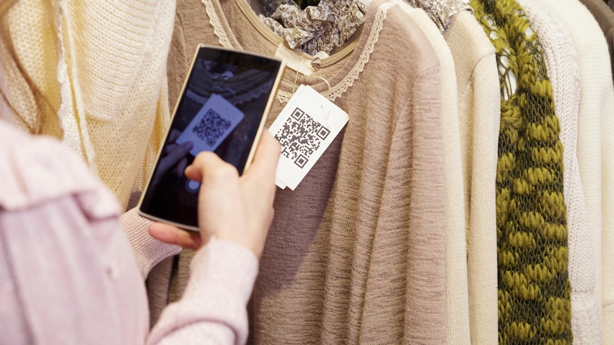 A hand holding a phone over a QR coded tag attached to a garment of clothing in a rack of garments