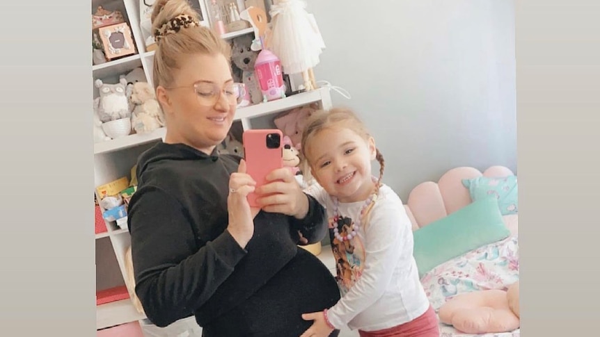 A pregnant woman taking a photo in a mirror while her young daughter holds her belly
