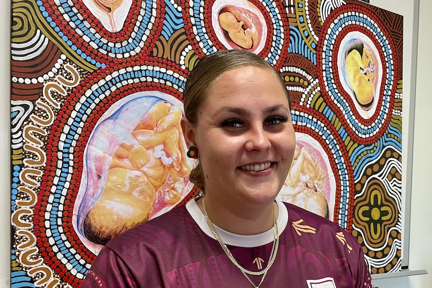 A portrait of a smiling woman in front of an Aboriginal artwork with babies in wombs