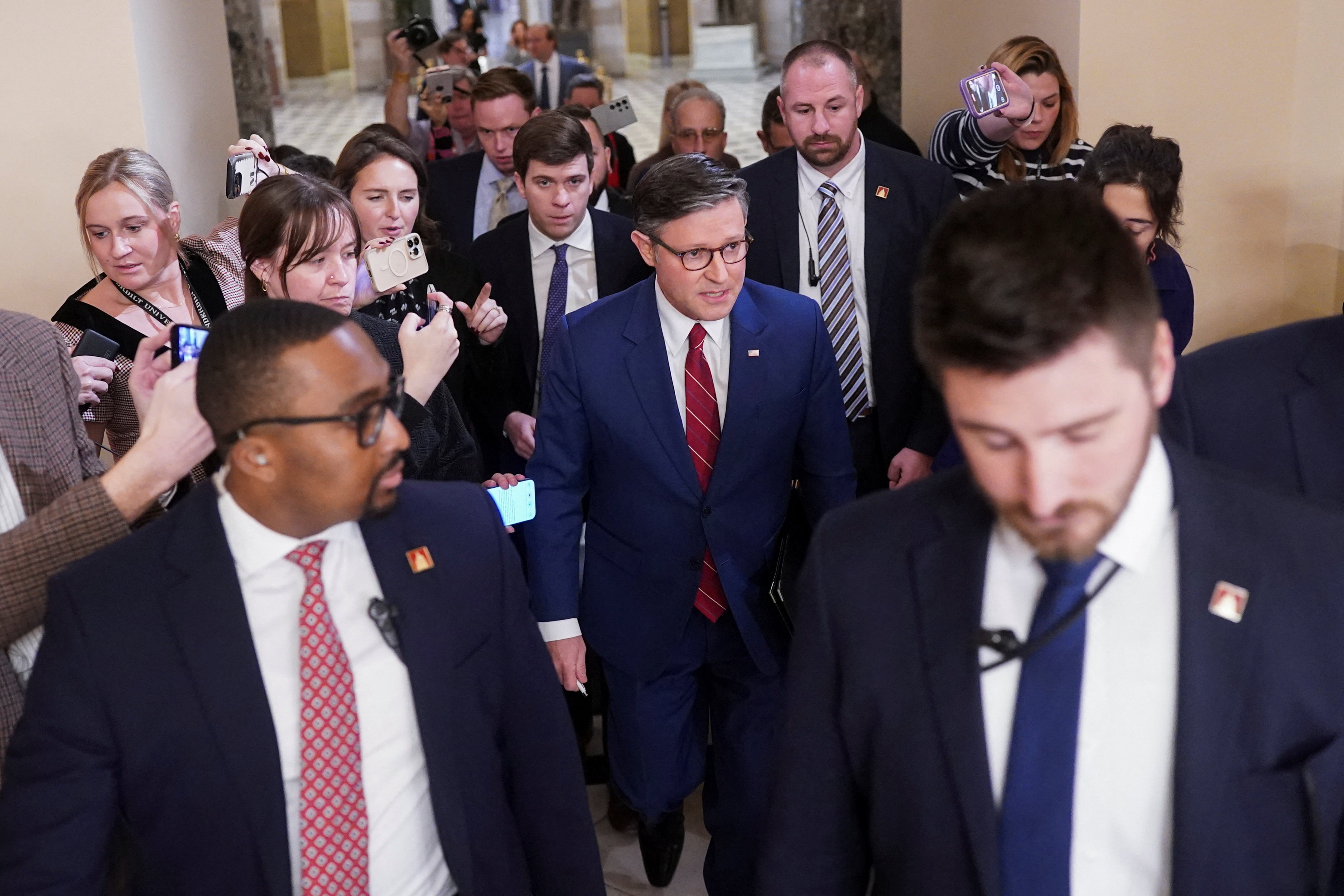 A crowd of people in suits walks through a hall surrounded by journalists with iphones