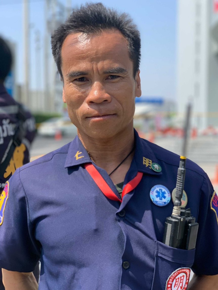 A close up of a Thai man with short hair wearing a blue uniform with a walkie talkie in his pocket.