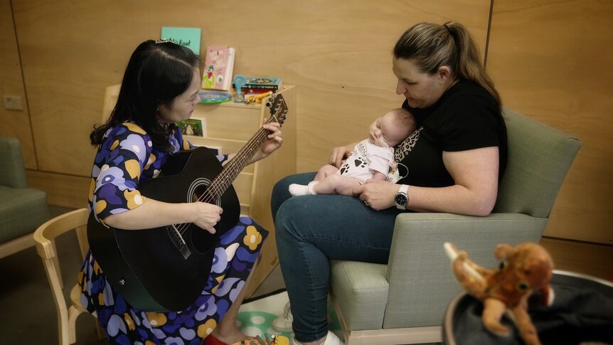 A woman plays the guitar to a mother holding her small baby in a chair