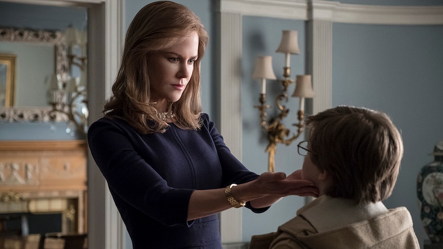 A well-dressed woman with pearl necklace holds a young boy's face in her hands in a simple ornately decorated powder blue room.