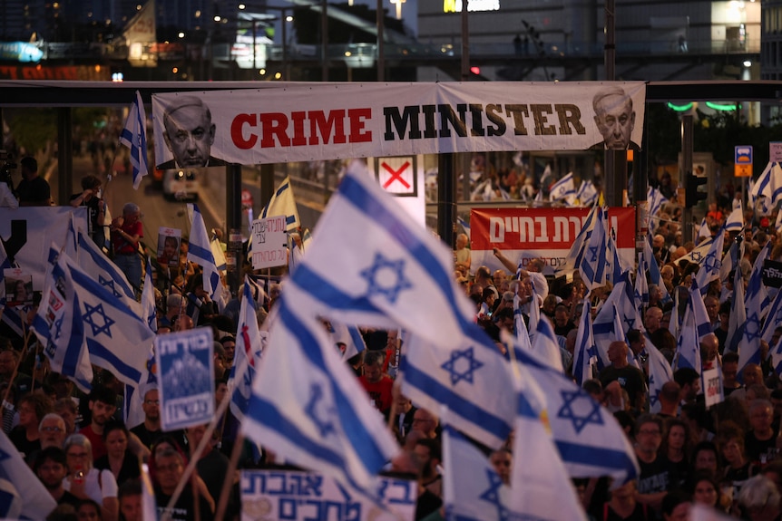 Crowd hold up Israeli flags in front of a banner that reads CRIME MINISTER with Netanyahu's face on it