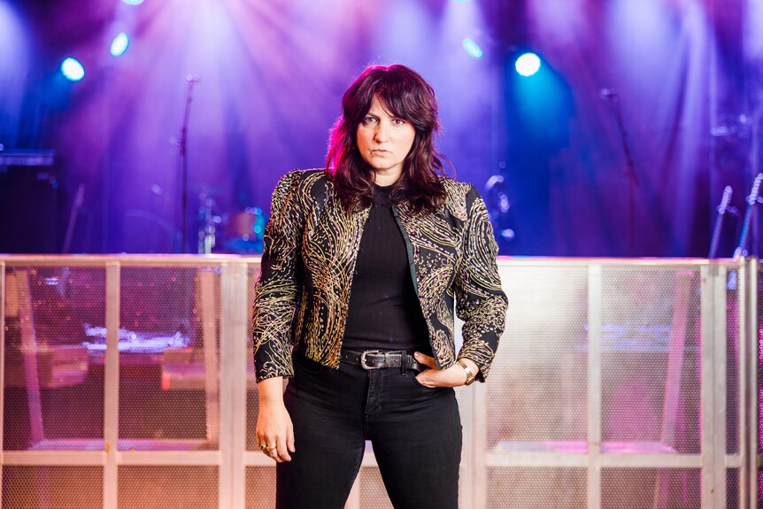 Tracy, wearing a gold and black jacket and black jeans, stands in front a stage that has bright blue lights