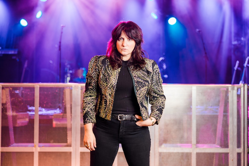 Tracy, wearing a gold and black jacket and black jeans, stands in front a stage that has bright blue lights