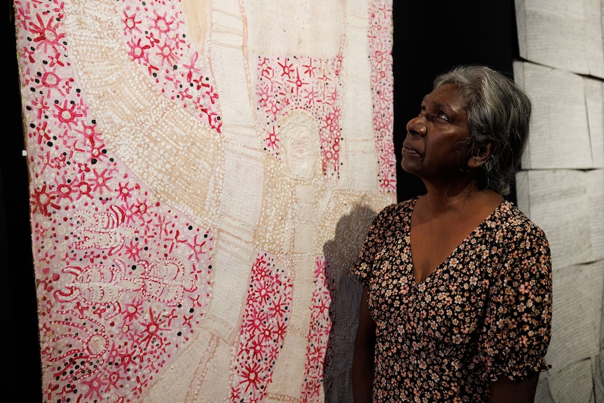 A woman stands next to a bark painting featuring cream-coloured water spirit figures and a red and pink sea.
