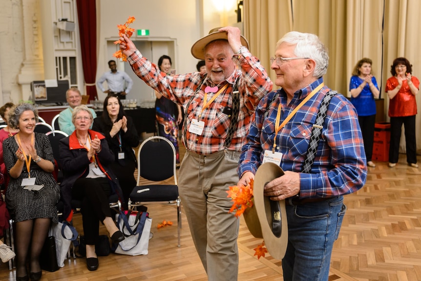 In a hall, two smiling men in flannelette shirts and trousers stand in front of a seated audience, who clap and smile.