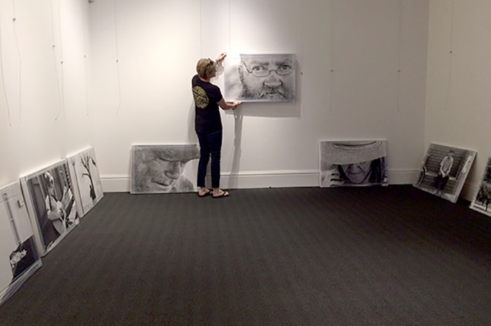 A woman hangs a blown-up black and white photograph of a man's face on a wall, with other photographs on the ground.