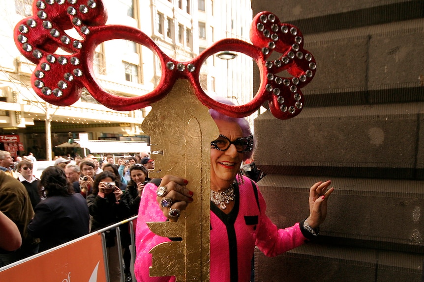 Barry Humphries is dressed as Dame Edna Everage as he holds a comically large key in the air, adorned with glasses.