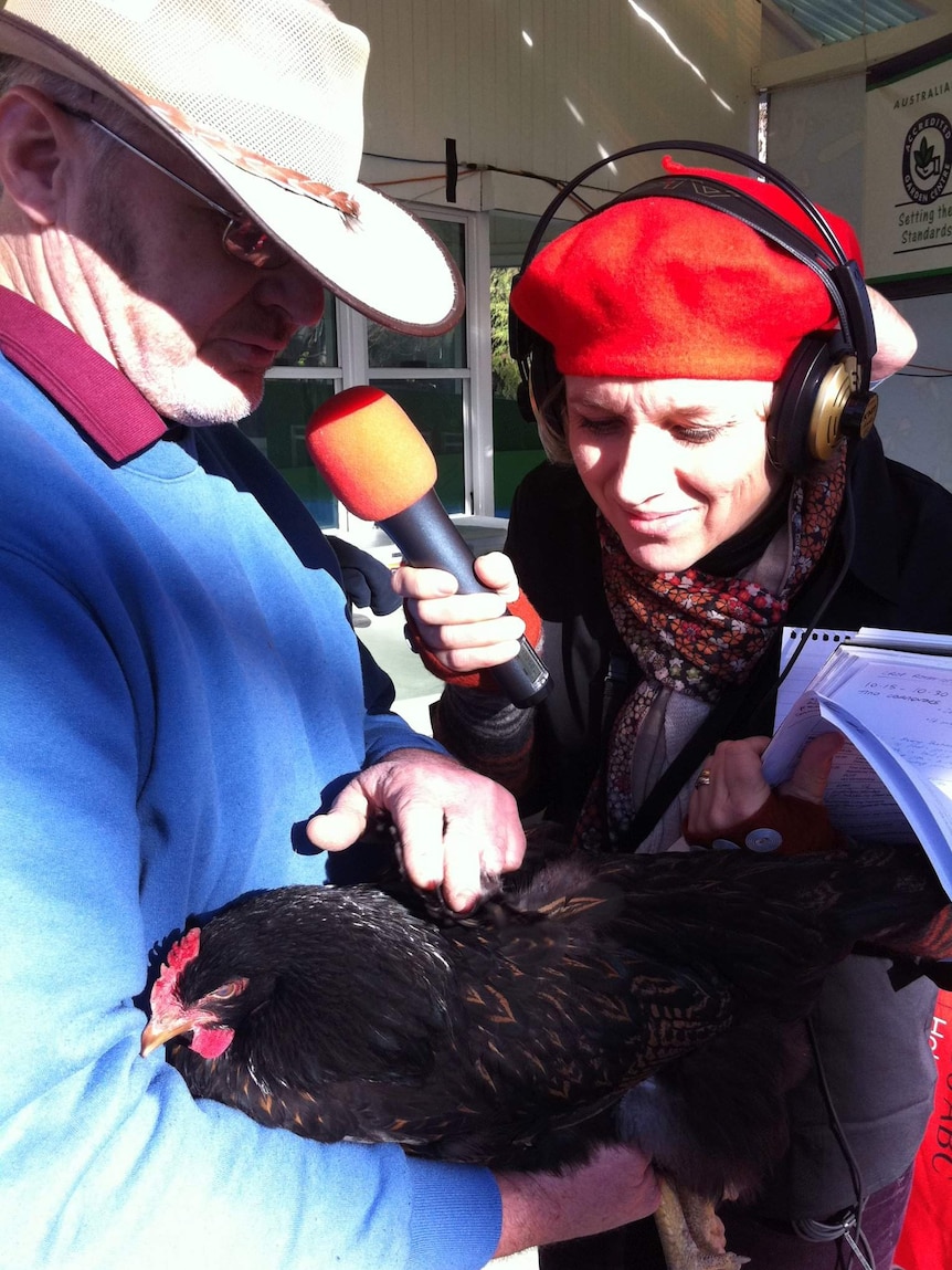 Jane is wearing a red beanie and large headphones and holding a microphone up to a man who is holding a chicken
