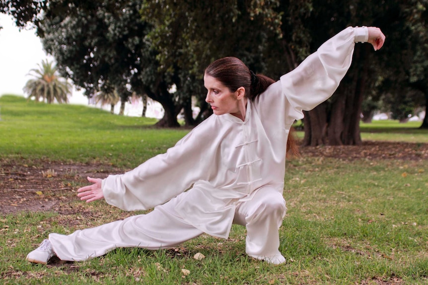 A woman lunges and holds her arms in the air, dressed in white.